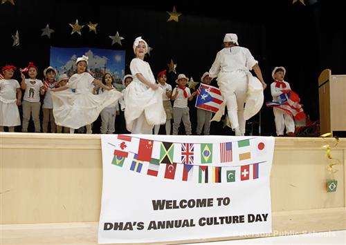 Young students parade on a stage that has a banner hanging on it that reads, "Welcome to DHA's Annual Culture Day".
